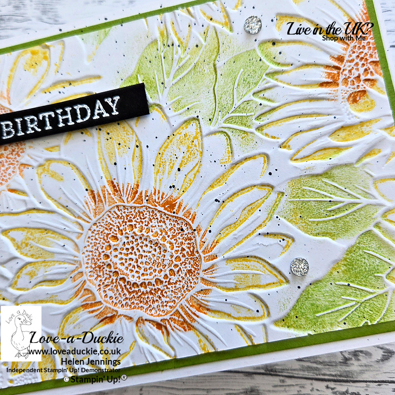 Close-up of a handmade sunflower birthday card featuring a letterpress technique using a 3D embossing folder. The embossed sunflower design is coloured with Daffodil Delight, Granny Apple Green and Pumpkin Pie inks on Basic White cardstock. A black heat-embossed “Happy Birthday” sentiment strip sits across the textured background with subtle black splatter and silver embellishments.